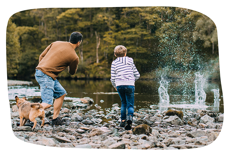 Ein Mann ist mit einem Jungen und einem Hund an einem Fluss oder See. Sie werfen zusammen Steine ins Wasser.